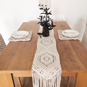May include: A white crocheted table runner with fringe on a wooden table. The runner has a diamond pattern and is centred on the table. There are two white placemats with white plates on them. A small plant in a glass vase is in the centre of the table runner.