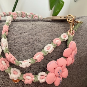 Puede incluir: Una correa de teléfono de crochet rosa y verde con flores de tela rosa. Las flores tienen una carita sonriente bordada.