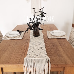 May include: A white macrame table runner with fringe on a wooden table. The runner has a diamond pattern and is centred on the table. A small vase with green plants is on the table runner.