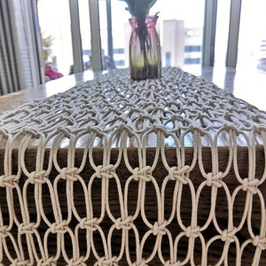 May include: A white crocheted table runner with a fringe edge. The runner is made of thick cotton cord and has a woven pattern.