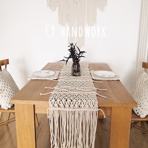 May include: A beige macrame table runner with fringe on a wooden table. The table runner is made with a knot pattern. There is a vase with a plant on the table. The text "CY HANDWORK" is visible on the wall behind the table.