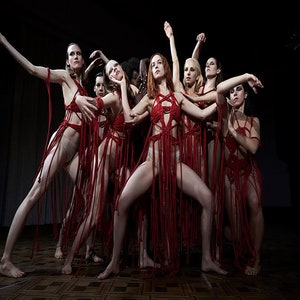 May include: A group of women wearing red, stringy outfits perform a dance on a stage. The outfits are made of red string and have a fringe-like appearance. The women are all in different poses, with their arms and legs extended.