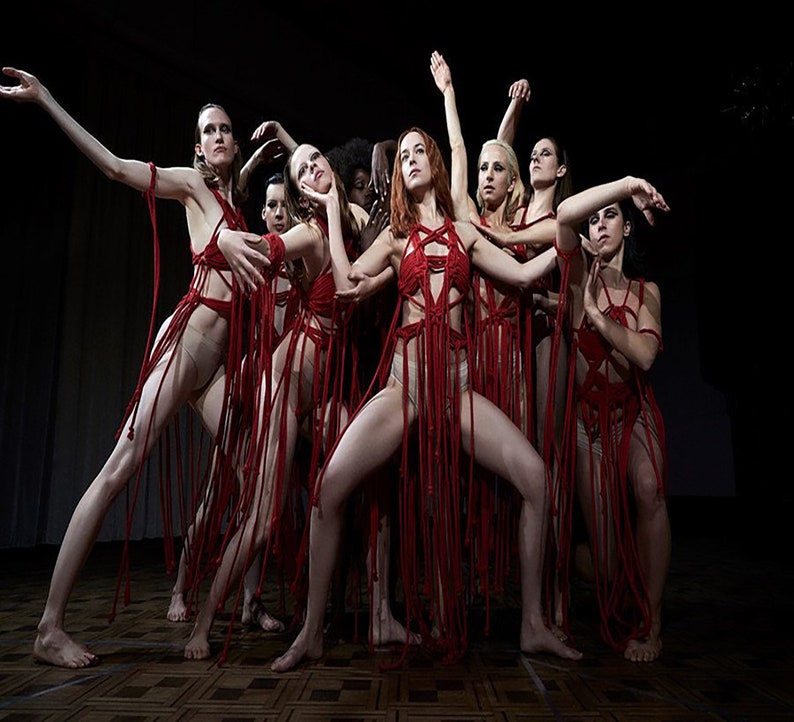 May include: A group of women wearing red, stringy outfits perform a dance on a stage. The outfits are made of red string and have a fringe-like appearance. The women are all in different poses, with their arms and legs extended.