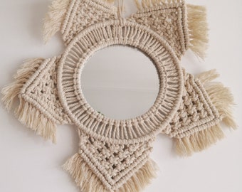 May include: A macrame mirror with a circular mirror in the center and a starburst design. The mirror is made of natural beige cotton rope with fringe details.