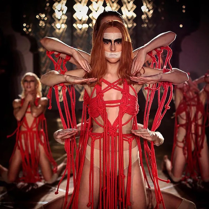 May include: Four people wearing red rope body suits with fringe. The person in the center has a white stripe across their eyes and mouth.