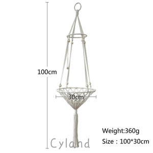 May include: White macrame plant hanger with a wooden bead detail. The hanger is 100 cm long and 30 cm wide.  The hanger has a tassel at the bottom.