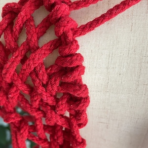 May include: Red macrame rope wall hanging with a knotted design. The rope is woven into a geometric pattern.