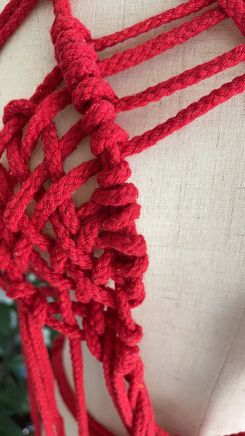 May include: Red macrame rope wall hanging with a knotted design. The rope is woven into a geometric pattern.
