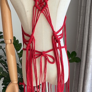 May include: Red macrame top with a criss-cross design and long fringe. The top is displayed on a white mannequin.