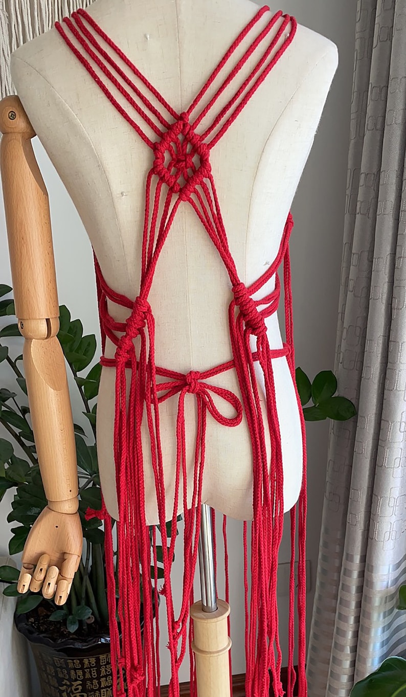 May include: Red macrame top with a criss-cross design and long fringe. The top is displayed on a white mannequin.