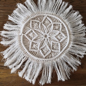 Puede incluir: Colgante de pared de macramé blanco con un diseño circular y flecos. El diseño presenta un patrón repetitivo de nudos y bucles.