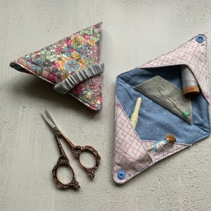 May include: Two fabric sewing project pouches. One is a triangular pouch with a floral print and a gray elastic band. The other is a folded pouch with a pink and white geometric pattern and a blue button. The pouch is open and shows a blue fabric with white dots, a gray fabric with pins, a spool of white thread, and a small glass bottle.