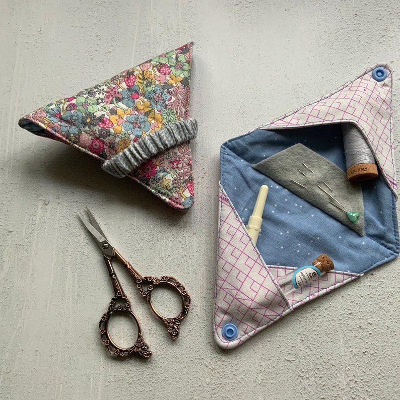 May include: Two fabric sewing project pouches. One is a triangular pouch with a floral print and a gray elastic band. The other is a folded pouch with a pink and white geometric pattern and a blue button. The pouch is open and shows a blue fabric with white dots, a gray fabric with pins, a spool of white thread, and a small glass bottle.