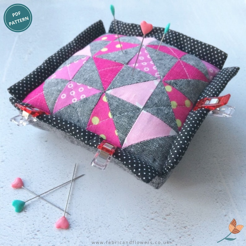 May include: A pink and gray patchwork pincushion with a black and white polka dot border. The pincushion is filled with pins and has a few red clips on the sides. The pincushion is sitting on a white surface.