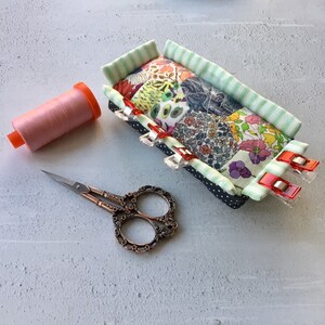 May include: A patchwork fabric pincushion with a green and white striped border. The pincushion is filled with pins and has four red and white clips attached to the sides. A spool of pink thread and a pair of ornate scissors are also in the image.