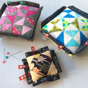 May include: Three fabric pincushions with different patterns. The first is pink, gray, and white with a polka dot pattern. The second is blue, green, and white with a geometric pattern. The third is black, yellow, and white with a musical instrument pattern. Each pincushion has a black border and is secured with red clips.