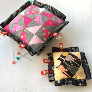 May include: Two fabric pincushions, one with a pink and gray geometric pattern and the other with a black and yellow guitar pattern. Both pincushions have black borders with white polka dots. The pincushions are sitting on a white surface.