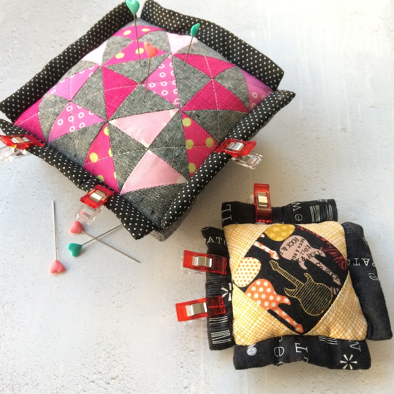 May include: Two fabric pincushions, one with a pink and gray geometric pattern and the other with a black and yellow guitar pattern. Both pincushions have black borders with white polka dots. The pincushions are sitting on a white surface.