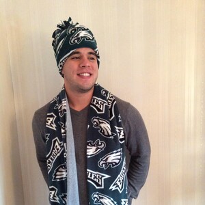 May include: A green and white fleece scarf featuring the Philadelphia Eagles logo. The scarf has a fringe on the ends and is being worn by someone with a matching green and white fleece hat.
