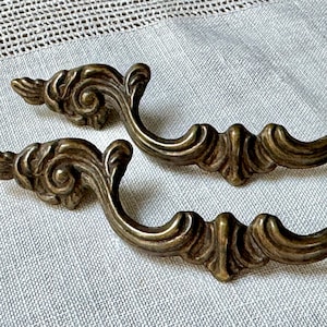 Pode incluir: Duas puxadores de gaveta de latão ornamentados com um design de rolos decorativos. As puxadores são de estilo vintage e têm um acabamento quente e envelhecido.
