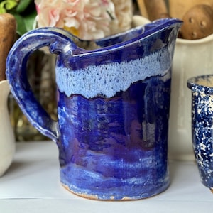 Könnte beinhalten: Handgefertigter Keramikkrug mit großem Henkel. Der Krug ist in Blautönen glasiert, mit einem helleren blauen Band um den oberen Rand. Der Krug wird wahrscheinlich zum Servieren von Getränken verwendet. Weitere Keramikartikel und Blumen sind im Hintergrund.