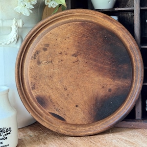May include: A round wooden cutting board with a dark brown finish, featuring a prominent raised outer rim and a centre surface marked by numerous visible scratches and signs of wear, showcasing a natural wood grain texture throughout the piece.