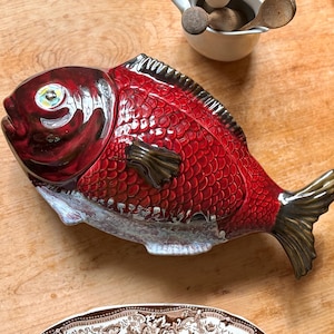 May include: A ceramic fish-shaped serving dish with a vibrant red glaze and detailed scale texture. The dish has a dark brown tail and fins, and a white and grey base. A decorative kitchen item.