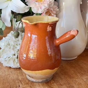 Puede incluir: Jarra de cerámica marrón con asa y borde amarillo. La jarra se encuentra sobre un fondo de flores y un jarrón de cerámica blanco. La jarra tiene un aspecto envejecido y desgastado.