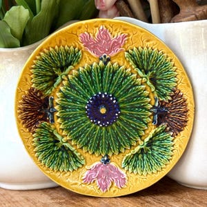 May include: A round ceramic plate featuring a yellow background with a central dark blue flower surrounded by green radiating leaves. The design includes four green leaf clusters, two pink floral motifs, and two brown textured elements.