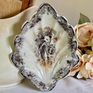 Może przedstawiać: Ozdobny talerz w kształcie liścia, biało-brązowy, z kwiatowym wzorem. Talerz ma falisty brzeg i centralną ilustrację kwiatów i postaci. Paleta kolorów obejmuje biel, brąz i beż.