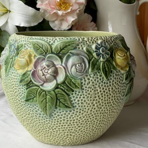 May include: A light green ceramic planter with a textured surface and embossed floral design. The planter features raised roses in shades of yellow, lavender, and white, surrounded by green leaves. The background is dotted, and the rim is a darker green.