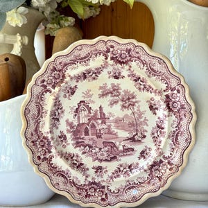 May include: A round ceramic plate with a scalloped edge featuring a deep purple transferware pattern on a cream background. The design depicts a landscape with architectural ruins, trees, and deer, surrounded by a floral and lattice border.