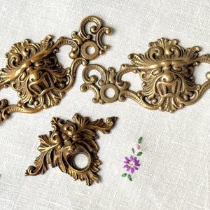 May include: Ornate brass drawer pulls and a single knob with a detailed face design. The hardware is a warm gold color, with intricate scrollwork and a vintage aesthetic. The pulls measure approximately 6 inches in length.