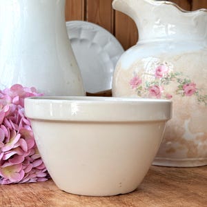 May include: A cream-colored ceramic bowl with a rounded shape, set against a backdrop of vintage pitchers and a plate. The bowl is the focal point, with a smooth, matte finish. Pink flowers add a touch of color to the scene, creating a rustic, farmhouse aesthetic.
