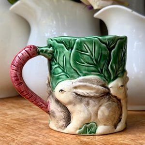 May include: A ceramic mug featuring a three-dimensional relief of rabbits nestled among green cabbage leaves. The handle is shaped like a textured, reddish-brown carrot. The mug has a glossy finish with visible fine crackle patterns throughout the glaze.
