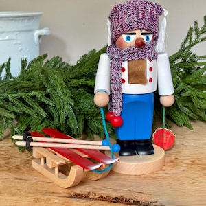 Könnte beinhalten: Eine Holz-Nussknacker-Figur, bekleidet mit einem weißen Hemd, einer blauen Hose und einem gemusterten Schal und Hut. Der Nussknacker hält einen roten Schmuck und steht neben einem Holzschlitten mit roten Skiern.
