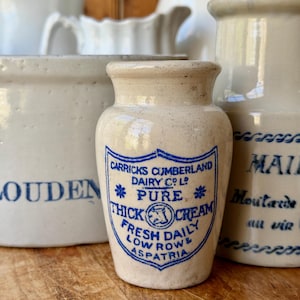 Puede incluir: Colección de tarros y jarras antiguos de cerámica. El tarro central es beige con una etiqueta azul que dice "Carricks Cumberland Dairy Co. Ltd. Pure Thick Cream". Otros tarros tienen letras azules. Los objetos están dispuestos sobre una superficie de madera.