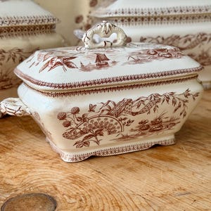 Peut inclure: Plat de service en céramique crème avec couvercle, orné d'un motif brun de paysages et de motifs floraux. Le plat est de forme rectangulaire avec une poignée décorative sur le couvercle et des poignées latérales. Le fond est une surface en bois.