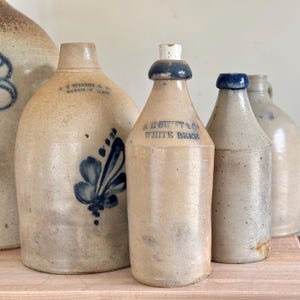 Op de afbeelding: Vier antieke steengoed flessen met blauwe en witte beschilderingen. De flessen zijn van verschillende groottes en vormen. Eén fles is gelabeld "M. Howitt & Co. White Beer".