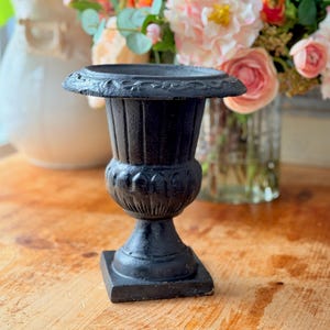 May include: A small, black, cast iron urn with a square base, a round body, and a decorative rim. The urn is displayed on a wooden surface, with a blurred background of flowers in vases.