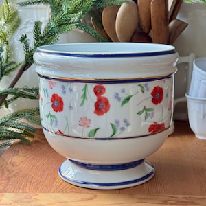 Op de afbeelding: Witte keramische plantenpot met een blauwe en gouden rand en een bloemenpatroon met rode klaprozen, roze bloemen en groene bladeren.