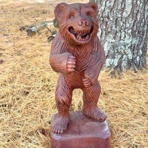 May include: A hand-carved wooden bear sculpture with a smiling expression. The bear is standing upright on a small, rectangular base. The sculpture is a reddish-brown colour and has detailed carvings of fur and facial features. The bear is outdoors, near a tree.