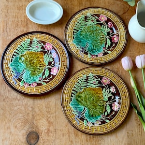 May include: Three round ceramic plates featuring a raised relief design of green fern leaves, large central leaves, and small pink flowers. The plates have a dark brown center and a wide border with a yellow and brown Greek key geometric pattern.