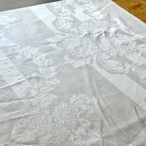 May include: A white and gray damask tablecloth with a floral and striped pattern. The fabric features large floral designs and vertical stripes, creating a classic and elegant look. The tablecloth is made of a soft, possibly linen, material.