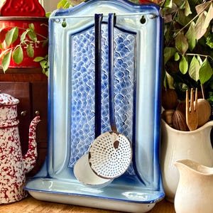 May include: A vintage blue and white enamel kitchen utensil holder with a matching skimmer and ladle. The holder has a decorative fish scale pattern and is mounted on a wooden surface. A red container and a patterned coffee pot are also visible.