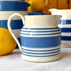 Puede incluir: Una jarra de cerámica blanca con rayas horizontales azules. La jarra tiene un asa curva y un pico para verter. Un limón es visible en primer plano, y otras jarras están en el fondo.