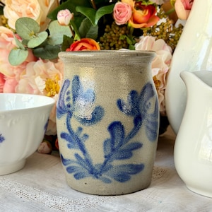 May include: A stoneware jar with a light beige background and blue floral designs. The jar is cylindrical with a slightly flared rim. Other white ceramic pieces and flowers are in the background.