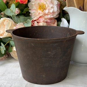 May include: A dark brown, weathered cast iron pot with a wide, open top and two small, flat side handles. The exterior surface shows a textured, oxidized patina with visible signs of age and wear, featuring a deep, rounded basin shape.