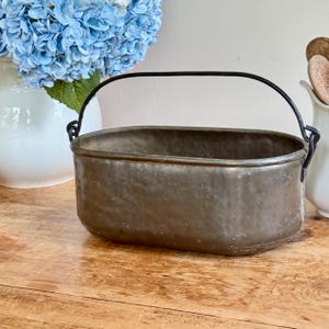 Puede incluir: Un cubo de metal ovalado con un asa negra. El cubo tiene un aspecto envejecido y desgastado, lo que sugiere que es antiguo. El asa se arquea sobre la parte superior, sujeta a los lados. El cubo está sobre una superficie de madera.