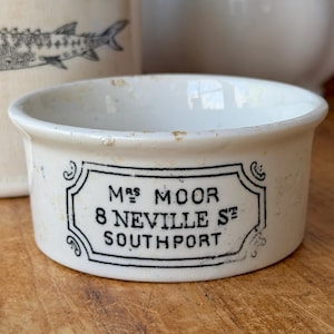 May include: A small, off-white ceramic dish with a black label that reads "MRS MOOR 8 NEVILLE ST SOUTHPORT". The dish has a rounded shape and a slightly worn appearance, suggesting age. Other ceramic items are in the background.